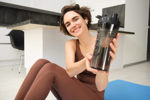 Smiling sportsgirl, woman after fitness training, gives you water bottle, stays hydrated after workout, sits on fitness yoga mat at home on floor.