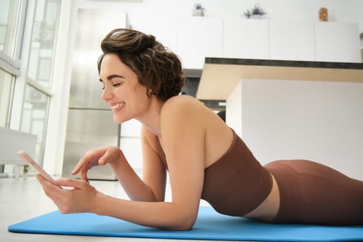 Fitness and technology. Smiling brunette woman in activewear, workout at home, looking at smartphone with happy face, watching training instructor on mobile phone app.