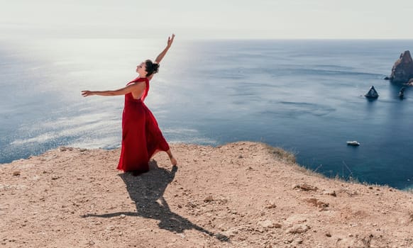 Side view a Young beautiful sensual woman in a red long dress posing on a rock high above the sea during sunrise. Girl on the nature on blue sky background. Fashion photo.