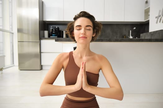 Portrait of female athlete, girl practice yoga, meditating in lotus pose, using mindfulness technique, workout at home on kitchen floor. Sport and wellbeing concept