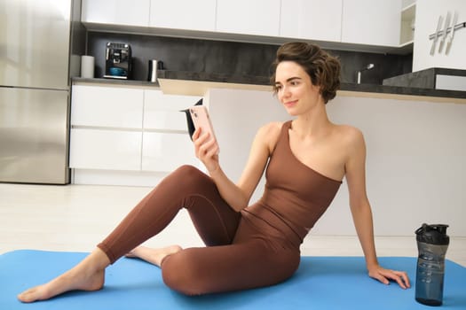 Young fitness instructor looking at her smartphone, sitting on rubber mat at home. Young woman doing workout training, looking at exercise videos on mobile app.