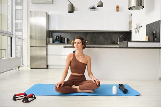 Portrait of young fitness woman, female athlete practice yoga from home, sitting in lotus pose with relaxed face, using rubber mat in kitchen for workout. Sport and wellbeing concept