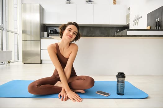 Portrait of fitness girl doing yoga on rubber mat at home, workout indoors in kitchen wearing sportswear, practice minfulness, sitting in lotus pose with water bottle next to her.