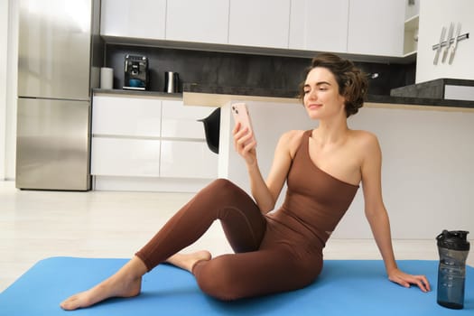 Portrait of young beautiful woman, fitness girl looking at her smartphone, taking break during workout at home in bright room. Sport and wellbeing concept