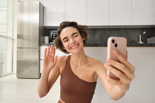 Image of young fitness girl blogger, records her workout at home, chats online while doing yoga in bright room. Woman with smartphone, joining remote yoga training via mobile phone.