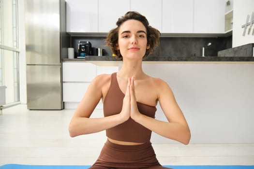 Close up portrait of young woman at home, sitting and meditating, practice yoga, making lotus pose, exercise indoors. Sport and women wellbeing concept