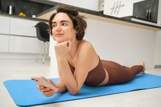 Online fitness and wellbeing. Portrait of young woman lying on yoga mat, looking at smartphone app, looking at workout tips, doing training at home in activewear.