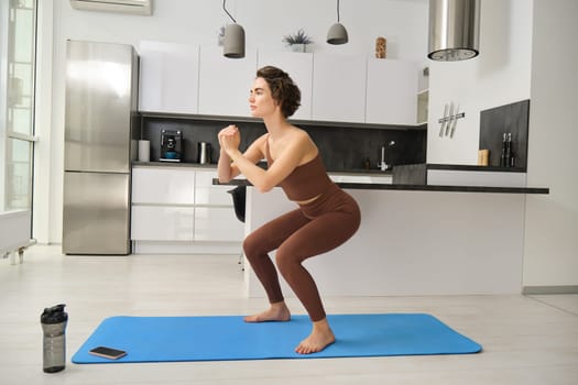 Gym at home. Young woman doing squats in bright room, workout indoors in sportswear, doing exercises on rubber yoga mat.