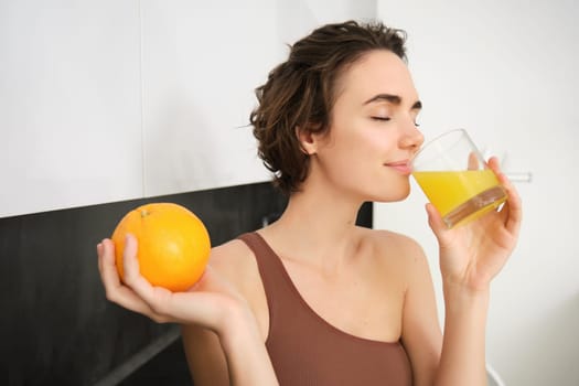 Image of sportswoman, fitness girl holding glass of juice and an orange, smiling, drinking vitamin beverage after workout, standing in her kitchen at home. Healthy lifestyle and sport concept.