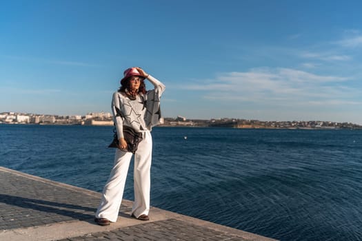 Stylish seashore woman. Fashionable woman in a hat, white trousers and a light sweater with a black pattern on the background of the sea