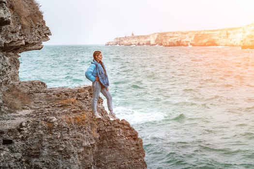 woman sea travel. A woman in a blue jacket stands on a rock above a cliff above the sea, looking at the stormy ocean. Girl traveler rests, thinks, dreams, enjoys nature.