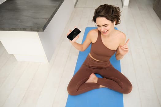 Dancing brunette girl sits on yoga mat, dances with headphones in ears, listens music during workout, sport training session from home, wears activewear, sits in yoga lotus pose.