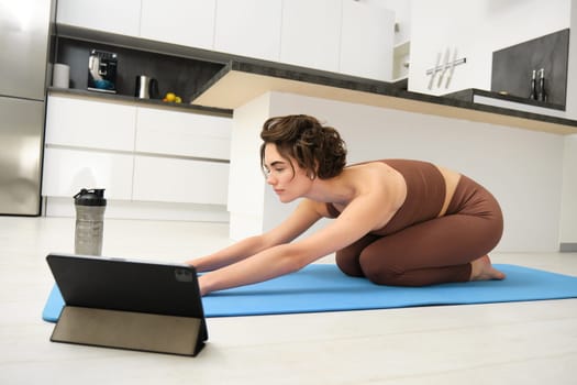 Image of young sportswoman does yoga at home, watches gym instructor tutorial video on tablet, stretches body, fitness training exercises, pilates class online from home.