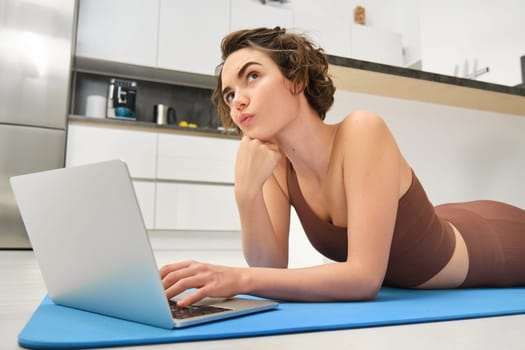 Portrait of young sportswoman, lying on floor at home, using yoga mat, workout, thinking while using laptop, searching for online gym exercises for training indoors. Sport and fitness concept
