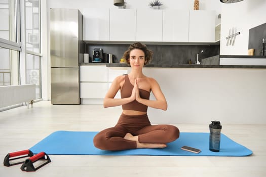 Portrait of female athlete, girl practice yoga, meditating in lotus pose, using mindfulness technique, workout at home on kitchen floor. Sport and wellbeing concept