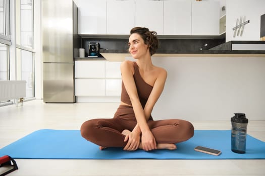Portrait of fitness girl doing yoga on rubber mat at home, workout indoors in kitchen wearing sportswear, practice minfulness, sitting in lotus pose with water bottle next to her.