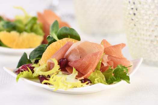 Portioned appetizer of prosciutto ham served on a of mixed with orange and pomegranate.