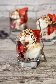 Summer cold dessert - Plombir ice cream scoop with chocolate sauce, cookie crumbs, ripe strawberries, crushed peanut nuts in a glass on a wooden table