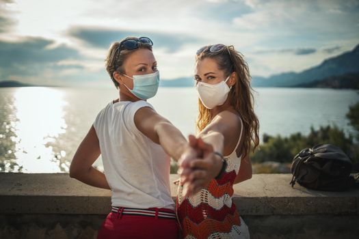 Shot of a happy female friends with protective mask spending time on seaside, enjoying in sunset, exploring a Mediterranean at corona pandemic.