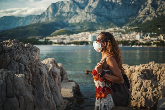 Smiling young female turist with protective mask spending time on seaside during exploring a Mediterranean at corona pandemic. 