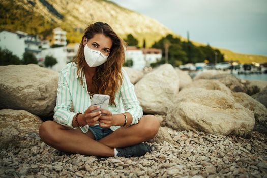 Shot of an attractive happy young woman wearing a protective N95 mask and using her smartphone while spending time on the seaside during the COVID-19.