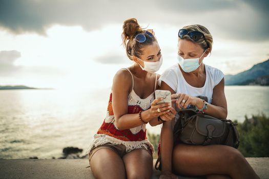 Shot of happy female friends with protective mask spending time on seaside and surfing social media during exploring a Mediterranean at corona pandemic. 