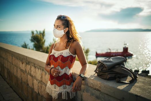 Shot of a happy young woman with protective mask spending time on seaside during exploring a Mediterranean at corona pandemic. 