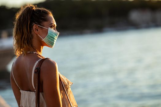 Shot of an attractive young woman wearing a surgical mask while enyoing herself during walk promenade by the sea.