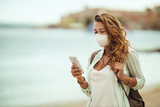 Shot of an attractive happy young woman wearing protective N95 mask and surfing social media on hers smartphone while enjoying a vacation on the beach during the COVID-19.