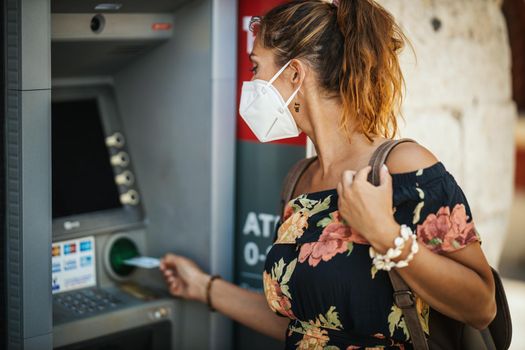 Side view of a young woman is wearing N95 protective mask while taking money from ATM machine during Covid-19 pandemic.