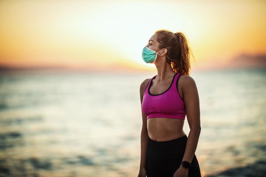 Shot of a female runner wearing protective face mask while doing training near the beach at sunset during the COVID-19 pandemic.