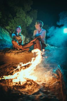 The happy smiling daughter and her mother are sitting on the sandy seashore by the fire in the evening.