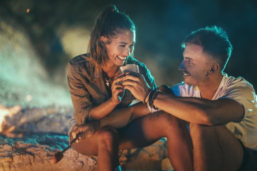 The happy smiling couple is sitting on the sandy seashore by the fire in the evening. They are frying sausages on stick.