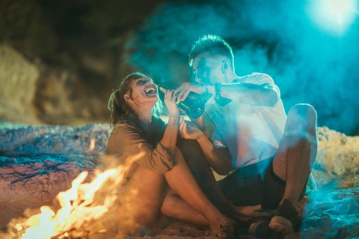 A happy family is sitting on the sandy seashore by the fire in the evening. They are making fun and drinking beer.