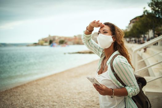 Shot of an attractive happy young woman wearing protective N95 mask and using smartphone while enjoying a vacation on the beach during the COVID-19.