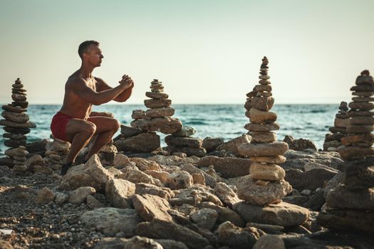 A handsome man is doing squat exercise at the sea beach in summer sunny day.