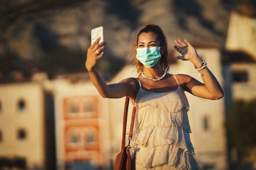 Shot of an attractive young woman wearing a surgical mask and making video call with her smartphone while exploring the wonderful city of Meditteranian.