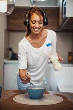 Beautiful young smiling woman is preparing her healthy breakfast in her kitchen and listening music.