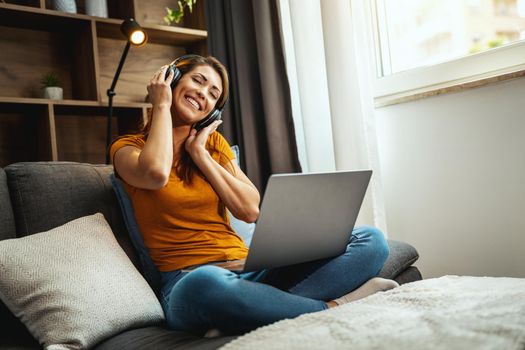 Shot of a young woman sitting cross legged on the sofa and using a laptop and headphones to listening music at home.