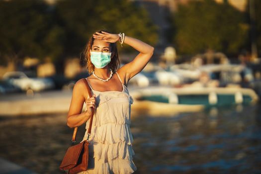 Shot of an attractive young woman wearing a surgical mask while enyoing herself during walk promenade by the sea.