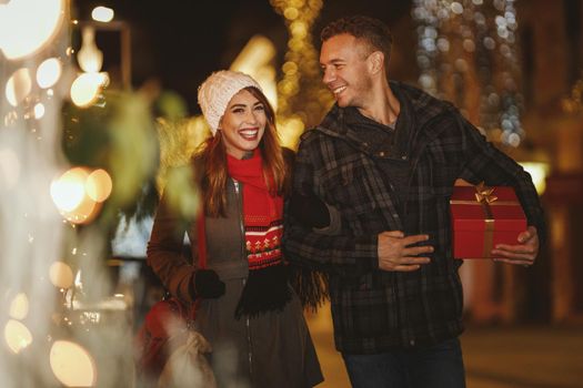 Shoot of a cheerful young couple are having fun in the city street at the Christmas night. They are laughing and buying presents for holidays to come.