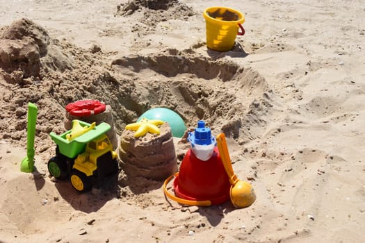 Children's plastic toys green end yellow car, shovel, yellow and red buckets, green ball with yellow sand on the beach by sea. Children's beach toys on sand on a sunny day. Sandbox on the playground