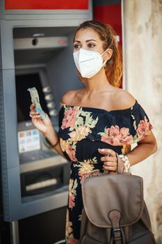 Side view of a young woman is wearing N95 protective mask while taking money from ATM machine during Covid-19 pandemic.