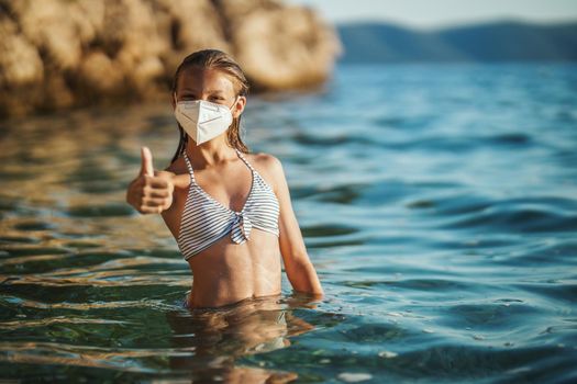 Shot of an happy teenager wearing surgical mask enjoying a vacation on the beach and showing thumb up during the COVID-19. Looking at camera.