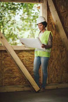 Shot of an African female architect checking plans at the construction site of a new wooden house. She is wearing protective workwear and white helmet.