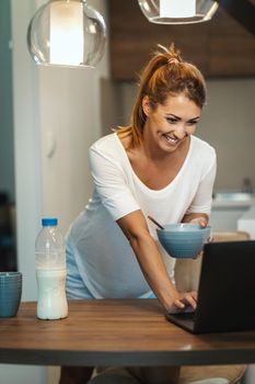 Beautiful young woman is eating her healthy breakfast in her kitchen and using laptop.