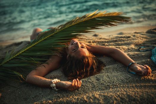 Shot of an attractive young woman is lying on the tropical sandy beach covered with palm tree leaf.