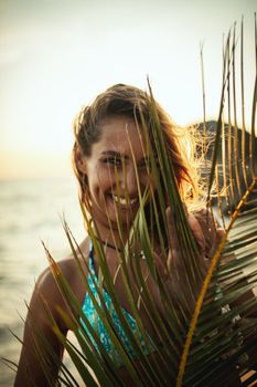 Portrait of a shy young woman is posing and looking under palm tree leaf at the tropical beach.
