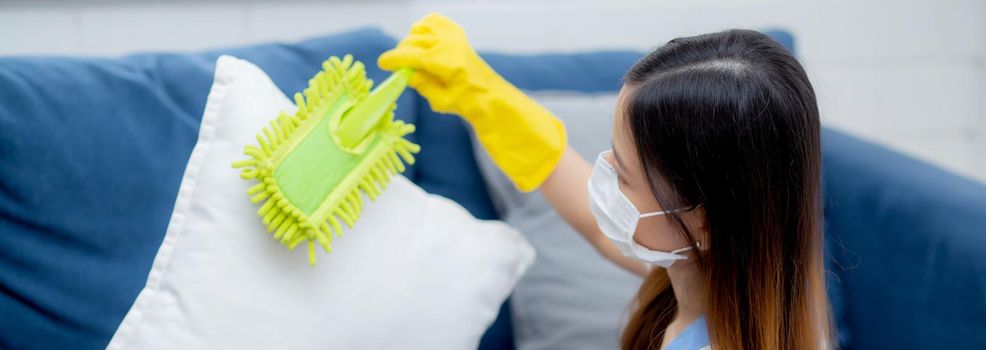 Young asian woman in face mask and gloves cleaning dust with duster on sofa and cushions at home, girl doing housework for hygiene at house, housekeeper and service, health care and lifestyle concept.