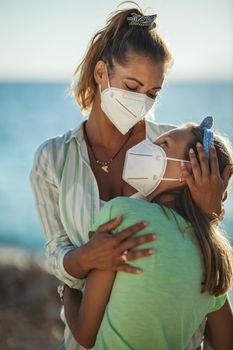 Shot of a happy little girl in her young mother's embrace with protective N95 mask spending time on the seashore at corona pandemic. 